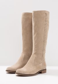 Bottes beige montant jusqu'au genou en daim avec un talon plat. Elles présentent un accent tressé le long du côté intérieur et une texture lisse.