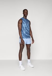 Blå tie-dye ærmeløs top, lyseblå shorts, hvide sneakers og sokker. Model står med hænderne afslappet ved siden af kroppen mod en neutral baggrund.