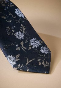 Cravate marine avec un motif floral en bleu clair et or, fabriquée en tissu texturé. L'extrémité effilée met en avant un détail de motif complexe.