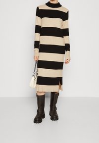 Randig klänning i beige och svart med ribbad krage och ärmslut, sidoklyfta, kombinerad med bruna knähöga stövlar och en liten vit väska.