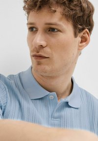 Svetlo modra polo majica z teksturiranim vzorcem in vertikalnimi črtami, kratkimi rokavi in ovratnikom s gumbom.