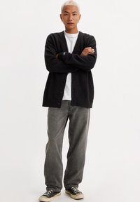 Black cardigan with a relaxed fit, button front, ribbed cuffs, and hem. Worn over a white t-shirt and paired with gray pants and black sneakers.