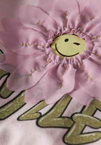 Tissu rose avec un grand motif de fleur comportant un visage souriant au centre. Accents en paillettes dorées sur le texte et les détails de la fleur avec des bords froncés.