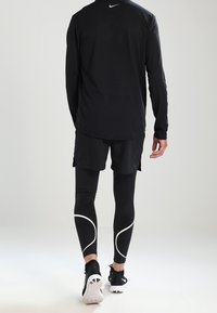 Schwarzes langärmliges Sportoberteil aus feuchtigkeitsableitendem Material mit Mesh-Details. Schwarze Shorts und eng anliegende Leggings mit weißen Akzenten.
