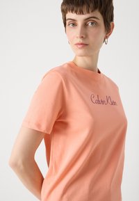 Persikinės spalvos medvilninė t-marškiniai su trumpomis rankovėmis, kuriuose krūtinėje violetine siuvinėta užrašas "Calvin Klein". Klasikinis apvalus kaklas.