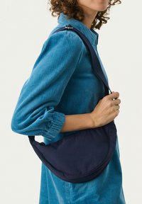 Borsa a spalla navy realizzata in tessuto morbido, caratterizzata da una forma a mezzaluna e una tracolla regolabile. Indossata sopra una camicia in corduroy blu texturizzata.