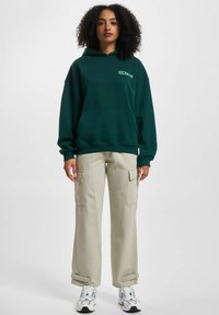 Green hoodie with front pocket and "ROCAWEAR" logo, paired with light beige cargo pants featuring side pockets and rolled cuffs.