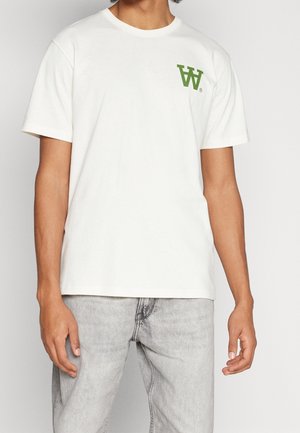 Mężczyzna w gładkim białym t-shircie z krótkim rękawem z zielonym logo "W" na lewej piersi oraz jasnoszarych dżinsach na białym tle.
