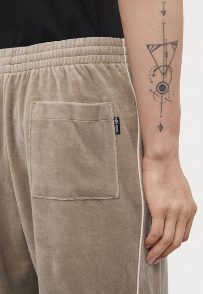 Beige corduroy joggers with a back pocket and elastic waist, featuring a white side stripe and a fabric label reading "NOON CLOODS."