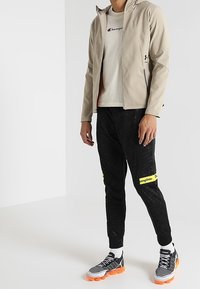 Ljusbeige zip-up jacka, vit t-shirt med logotyp, svarta sweatpants med gula detaljer och gråsvarta träningsskor med orange sulor.