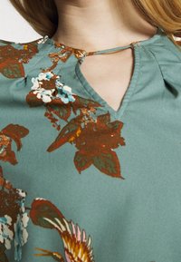 Hedvábná blůza s květinovým vzorem v barvě tyrkysové s sklady, zdobená oranžovými a bílými květy a výstřihem ve tvaru klíčové dírky. Hladká textilie.