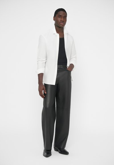 Chemise blanche à manches longues avec boutons portée sur un débardeur noir, associée à un pantalon large en cuir noir et des chaussures noires. Design simple et élégant.