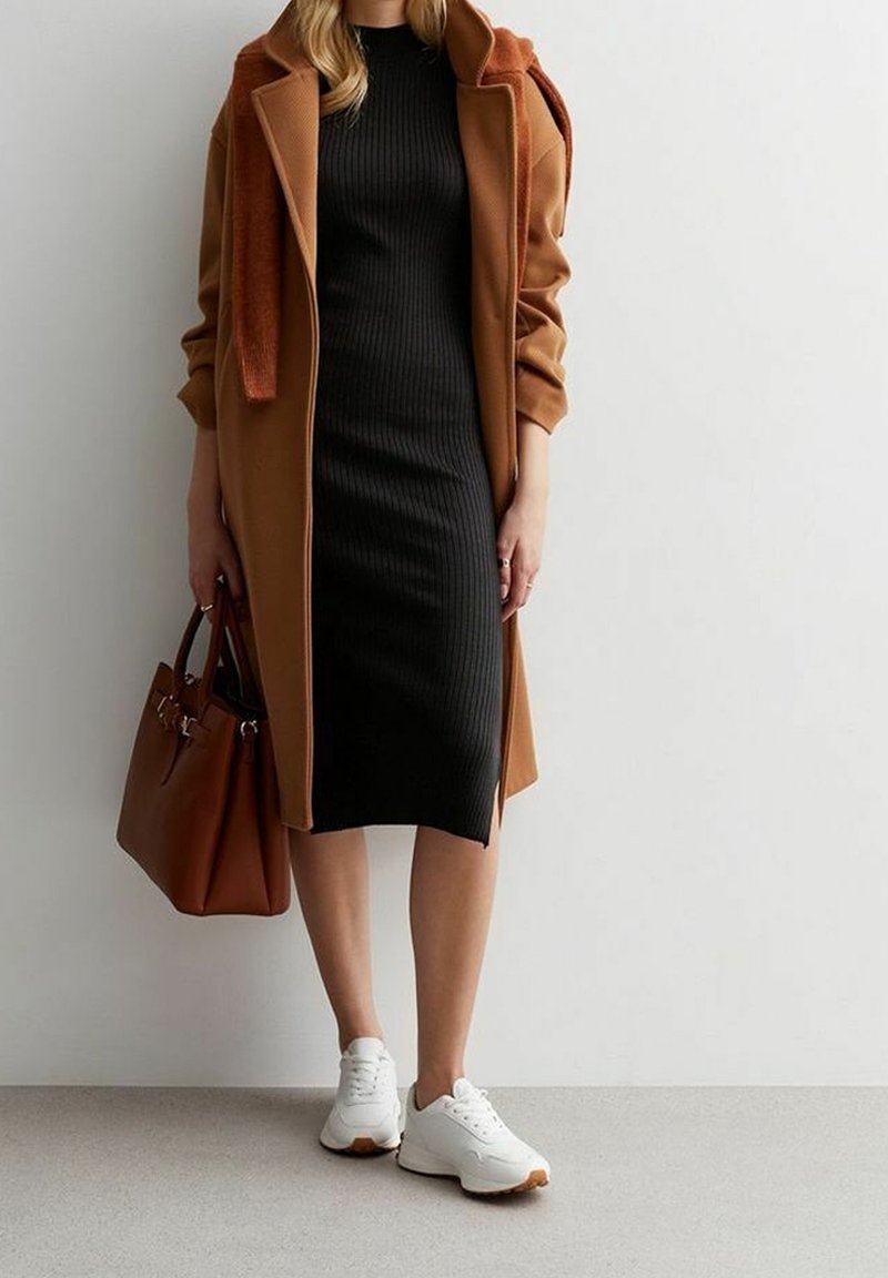 Manteau marron, robe noire à côtes, baskets blanches avec semelles beiges, sac à main marron. L'ensemble présente des textures superposées et des couleurs contrastées.