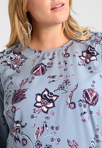 Blusa de color azul claro con estampado floral en azul marino oscuro y burdeos. Presenta un escote redondo y mangas cortas, con una textura suave y lisa.