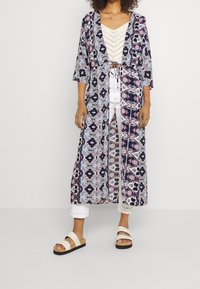 Lange bedruckte Kimono-Jacke mit einem Muster in Blau, Weiß und Pink, mit weiten Ärmeln und offener Front. Kombiniert mit einem crème-farbenen Oberteil und weißen Hosen.