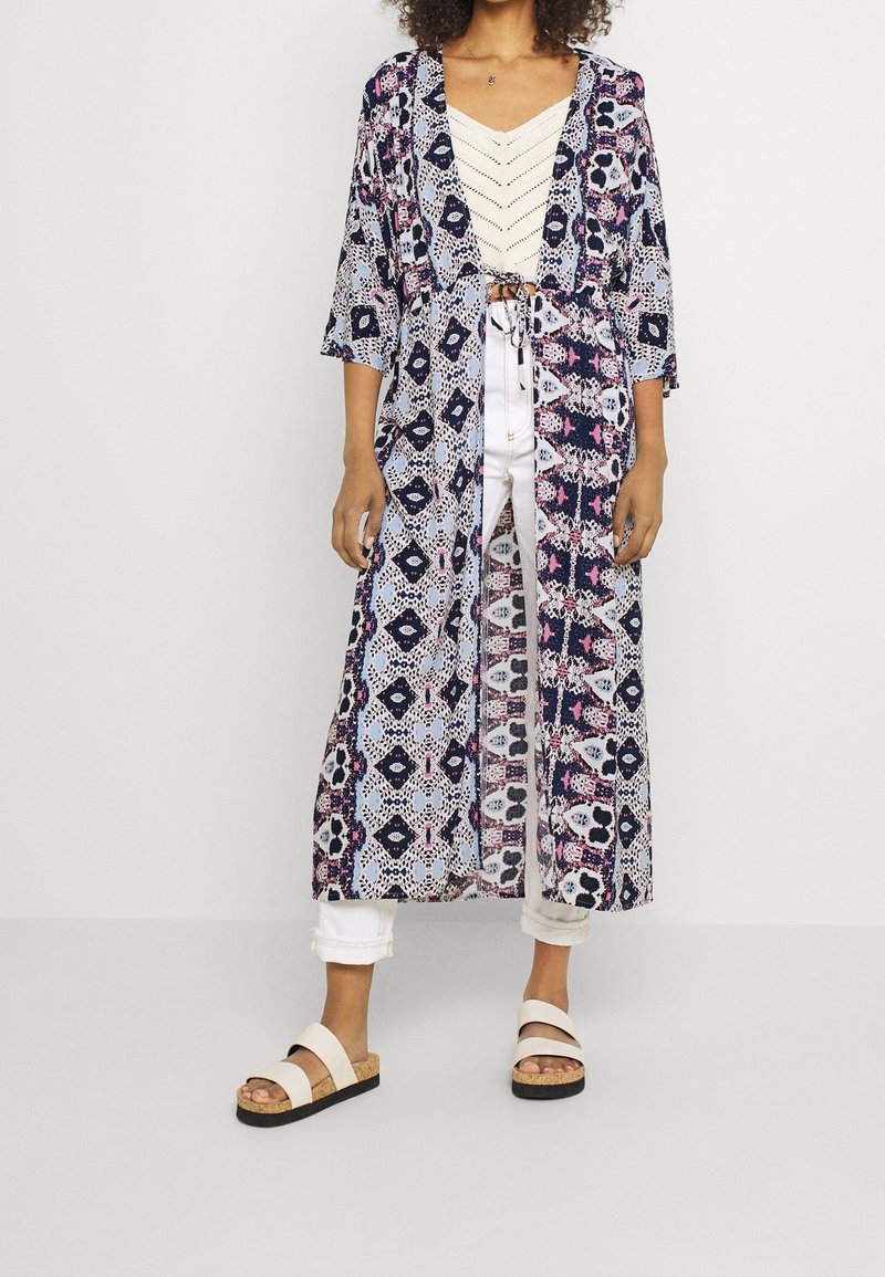 Lange bedruckte Kimono-Jacke mit einem Muster in Blau, Weiß und Pink, mit weiten Ärmeln und offener Front. Kombiniert mit einem crème-farbenen Oberteil und weißen Hosen.