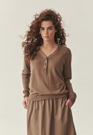 Žena s kučeravými vlasmi, oblečená v hnedom svetríku na gombíky s dlhým rukávom a kratšej sukni rovnakej farby, stojaca s rukami v vreckách na jednoliatom pozadí.