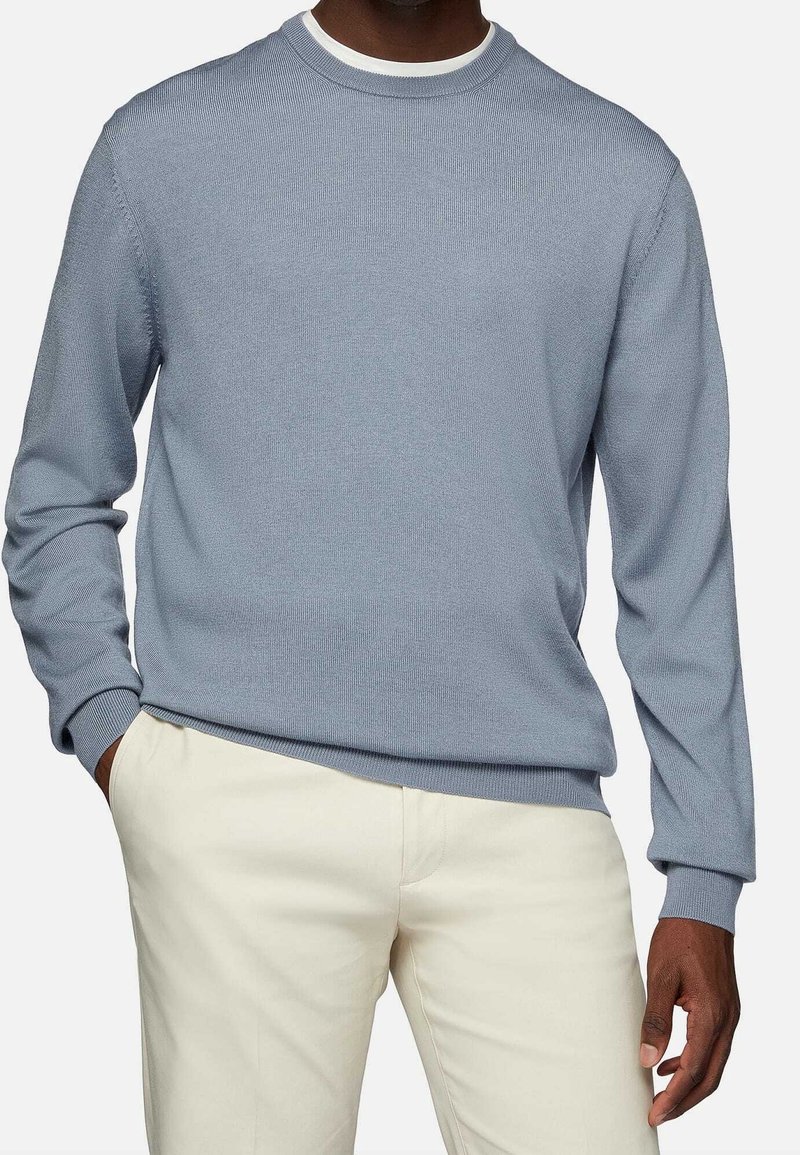Homme portant un pull bleu clair à col rond sur une chemise blanche avec un pantalon beige, mains dans les poches, sur fond uni.