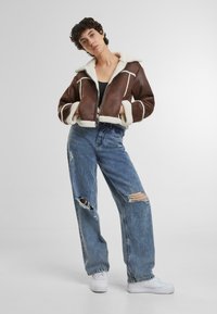 Giacca in shearling marrone con accenti bianchi, chiusura con zip e colletto ampio, abbinata a jeans azzurri chiari strappati e sneakers bianche.