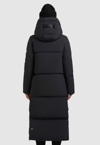 Persona che indossa un lungo cappotto invernale imbottito nero e un berretto nero, rivolto di spalle, in piedi contro uno sfondo semplice grigio chiaro.