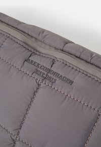 Tessuto grigio trapuntato con branding in rilievo "ARKK COPENHAGEN EST. 2015", cuciture scure e una finitura liscia e lucida.