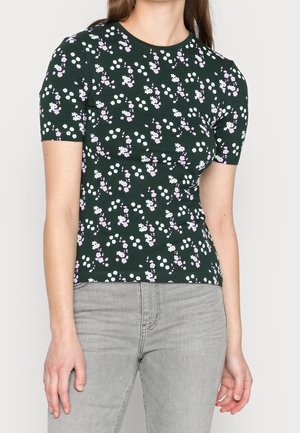 Frau trägt ein dunkelgrünes, eng anliegendes Kurzarmoberteil mit kleinen weißen und hellvioletten Blumenmustern, kombiniert mit hellgrauen Jeans.