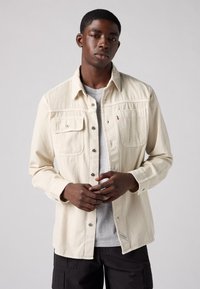 Beige button-up shirt with two chest pockets, made of a textured fabric, featuring metal buttons and a side label. Worn over a gray tee.