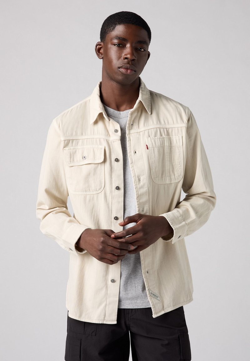 Beige button-up shirt with two chest pockets, made of a textured fabric, featuring metal buttons and a side label. Worn over a gray tee.