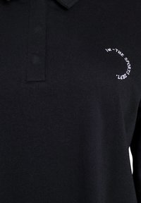 Schwarzes Poloshirt mit kurzen Ärmeln, aus einem weichen Stoff gefertigt. Verfügt über einen Kragen und ein weißes Logodesign auf der Brust.