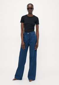 Zwarte fitted t-shirt in combinatie met hoge taille, wijde denim jeans in blauw. Het model draagt donkere zonnebrillen en schoenen met een opvallend patroon.