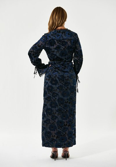 Robe longue en dentelle florale bleu marine avec une taille à nouer et des manches fluides. Présente une superposition transparente et une texture motif. Chaussures à talons hauts visibles.