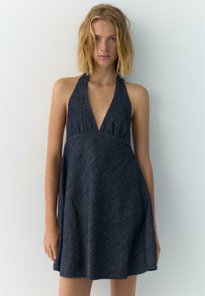 Młoda kobieta z blond włosami do ramion, ubrana w ciemną, bezrękawnikową sukienkę jeansową na jednolitym białym tle.