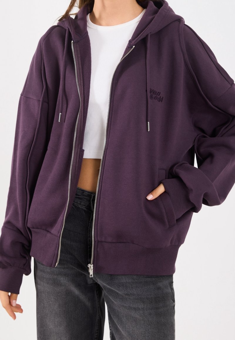 Personne portant un sweat à capuche violet ouvert par-dessus un crop top blanc et un jean gris foncé, avec une main dans la poche du sweat.