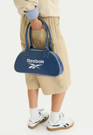 Persona che indossa pantaloncini beige e sneakers bianche Reebok, che tiene una borsa Reebok blu navy con logo bianco dalle maniglie.