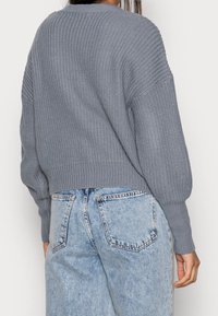 Szary sweter z dzianiny o luźnym kroju, z prążkowanymi mankietami i dołem. Połączony z jasnoniebieskimi dżinsami o wysokim stanie, pokazując widok z tyłu.