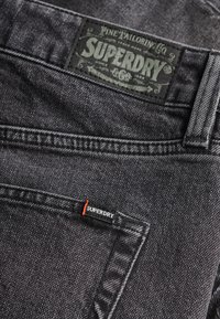 Jeans en denim noir avec une pièce vintage portant la marque "SUPERDRY" et une petite étiquette avec un accent rouge sur la poche avant.