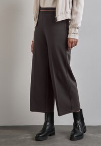 Pantaloni a gamba ampia di colore grigio scuro, realizzati in un tessuto morbido, con una superficie liscia e un elastico in vita, abbinati a stivaletti neri alla caviglia.