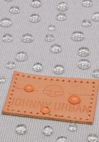 Vízlepergető szürke szövet, rajta tiszta vízcseppek; a jobb alsó sarokban "JOHNNY URBAN" felirattal domborított barna bőr címke.