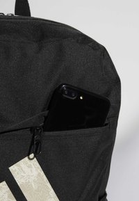Crni platneni ruksak s teksturiranom površinom, s bočnom džepom u kojem se nalazi crni pametni telefon i bež detaljem s logotipom.