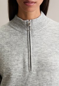 Grau melierter Pullover mit Reißverschluss und hohem Kragen, aus weichem Stoff gefertigt, mit silbernem Reißverschluss-Detail und strukturiertem, meliertem Oberflächenbereich.