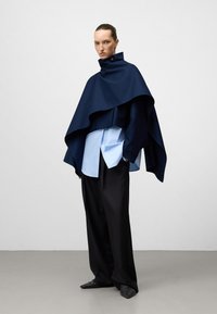 Cappotto navy con colletto alto sovrapposto a una camicia leggera azzurra con bottoni, abbinato a pantaloni neri larghi e scarpe nere texturizzate.