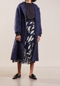 Donna che indossa un lungo cappotto peloso blu navy, una blusa nera con volant, una gonna plissettata con stampa astratta e mocassini neri lucidi, mentre si trova su un pavimento beige.
