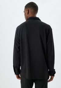 Schwarzes Langarmshirt aus glattem Stoff mit lockerer Passform, ausgestattet mit einem umlegbaren Kragen und leicht hochgerollten Bündchen.