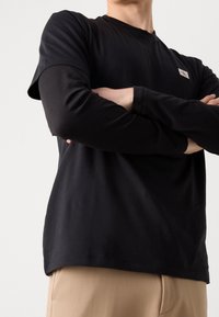 Czarny longsleeve z okrągłym dekoltem i małą naszywką logo, zestawiony ze jasnobeżowymi spodniami dla kontrastu.