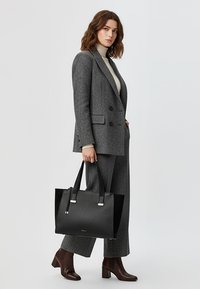 Frau trägt ein grau gemustertes Sakko und Hose, cremefarbenen Rollkragenpullover, braune Stiefeletten, hält eine große schwarze Handtasche und steht vor weißem Hintergrund.