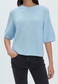 Femme portant un pull court à manches courtes tricoté bleu clair avec un pantalon noir, une main dans la poche, sur un fond uni.