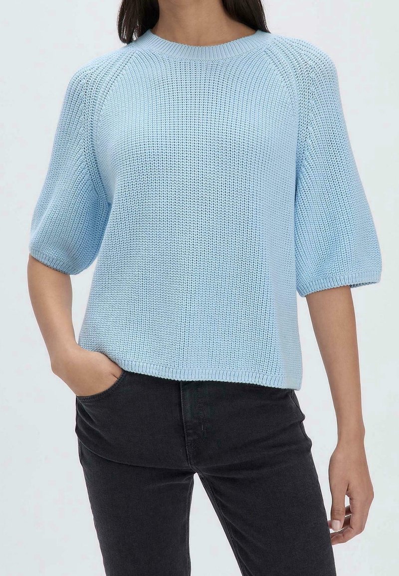 Femme portant un pull court à manches courtes tricoté bleu clair avec un pantalon noir, une main dans la poche, sur un fond uni.