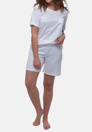 Kvinde med krøllet hår iført en hvid kortærmet skjorte og matchende hvide shorts, står barfodet mod en ensfarvet baggrund.