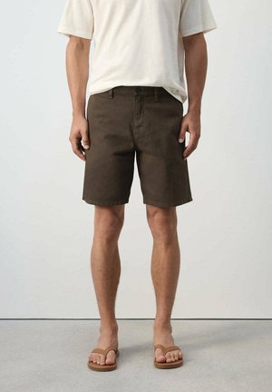 Hombre con pantalones cortos marrón oscuro, camisa blanca de manga corta y sandalias beige, de pie frente a un fondo liso.