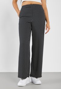 Black wide-leg pants with white polka dots, featuring a high waistband and side pockets, paired with white sneakers on a gray floor.